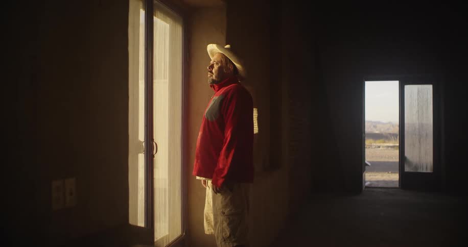 Hispanic man wearing a hat looks outside a window at home at morning