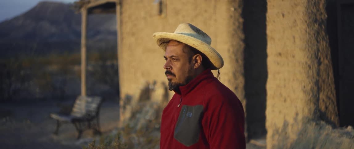 A hispanic older male standing outside near a small house and mountains