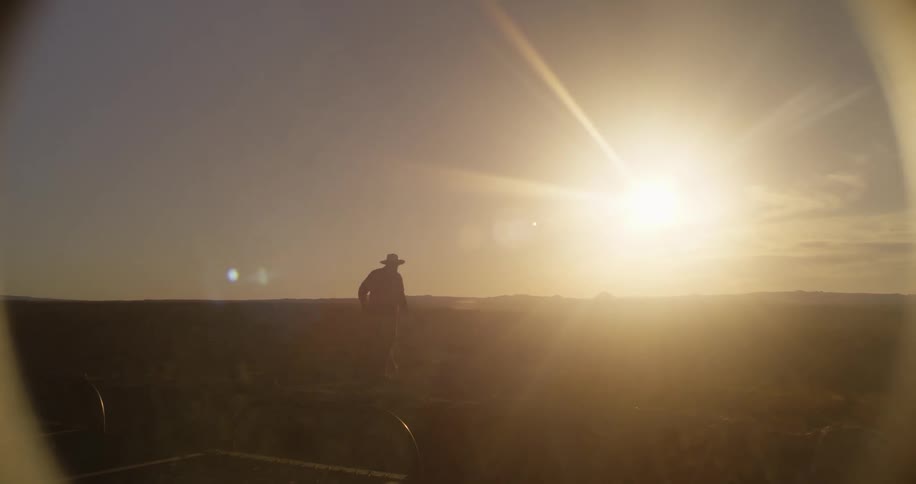 A silhouette of a man with a hat walking in a grassy landscape with the sun sky and clouds in the background