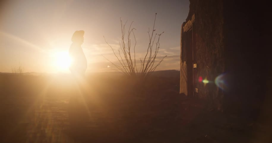 A male in a cowboy hat standing in sun flare in a desert near a small building