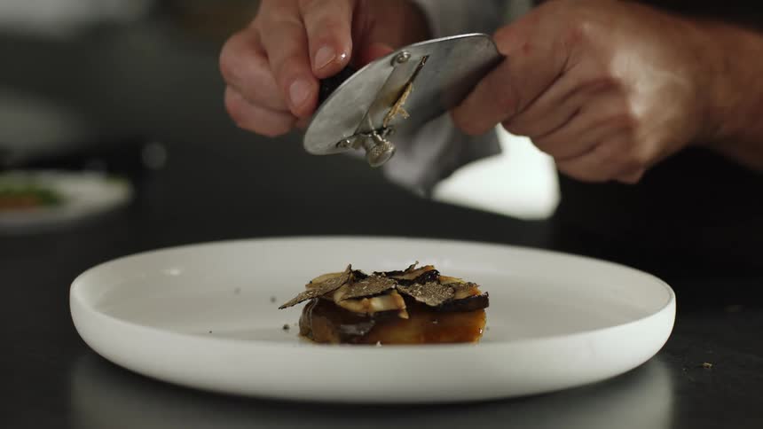 A chef uses a mandolin to drop sliced mushrooms onto an entree
