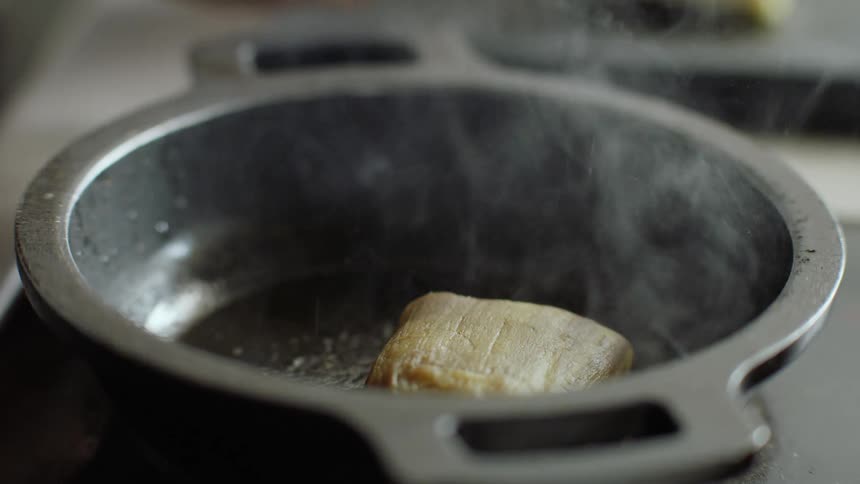 A cook is placing square shaped meat into a pan and smoke rises