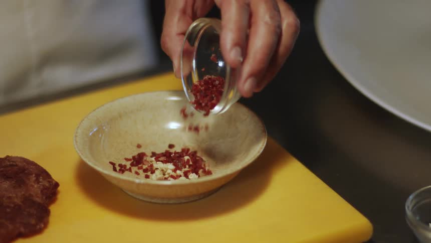 A hand reaches for a small glass bowl of spice and adds it to a bowl of chopped garlic