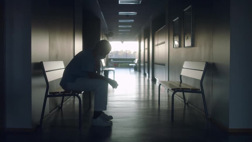 Nurse sits on bench in empty hospital corridor looking down with window showing outdoors in distance