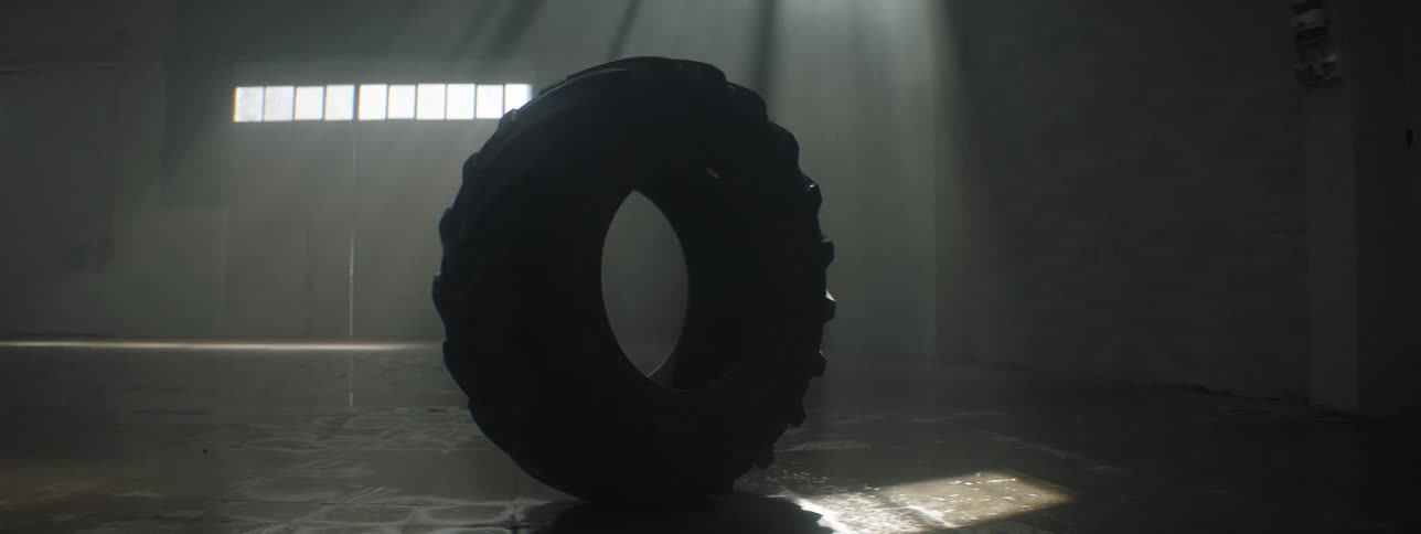 A large truck tire is standing by itself on a garage floor as light from the windows shines on it
