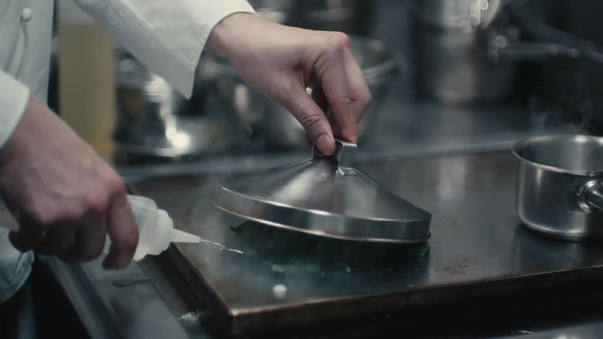 A chef stands at an industrial sized oven and squirts liquid on a burner and holds a pot lid above it to create steam