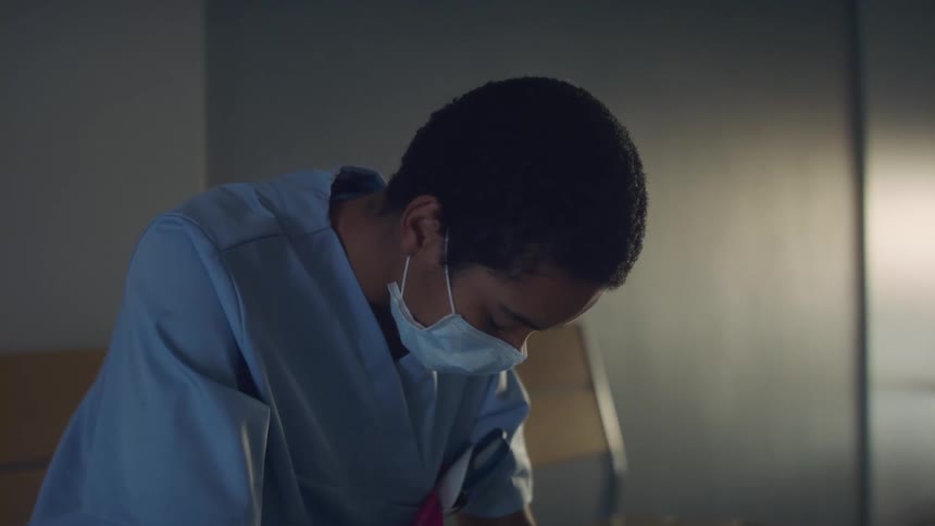 Young nurse wearing a blue disposable face mask sits tired inside a room