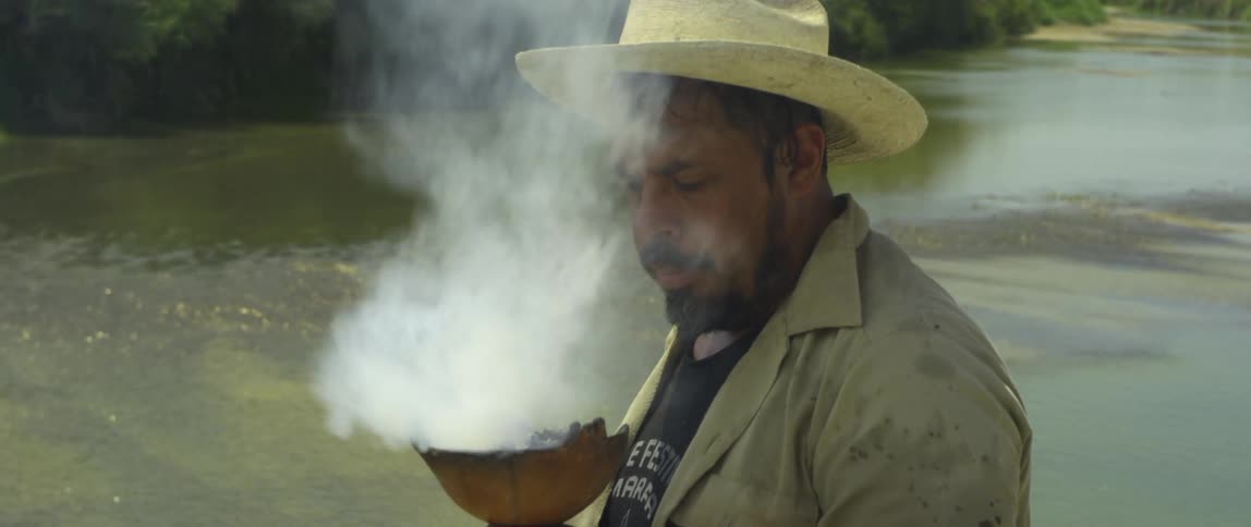 Man wearing a hat blows to stokes a fire inside a wooden bowl near a river