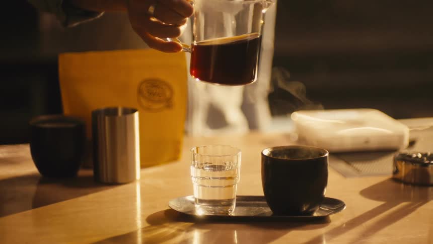 Someone is holding a pot of coffee and pouring it into a cup on a table