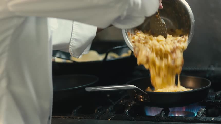 Chef wearing white stirs mixture before pouring it into skillet over burning gas stove