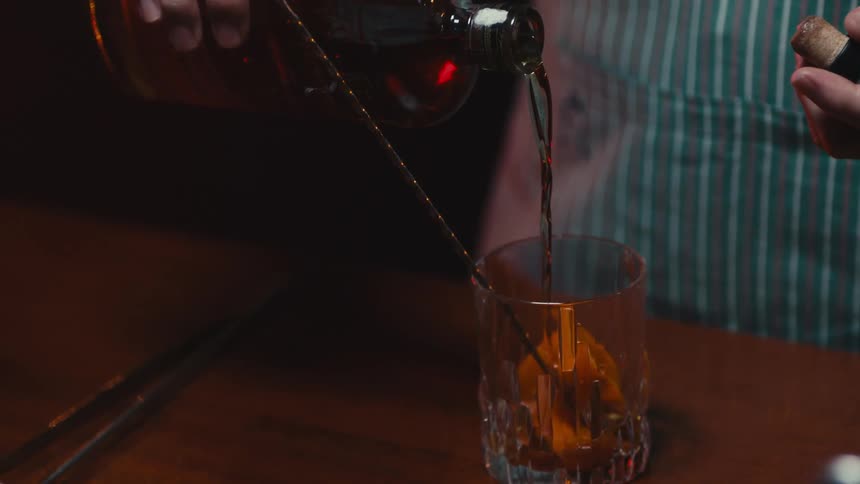 A bartender is pouring an alcoholic beverage into a glass tumbler in a bar