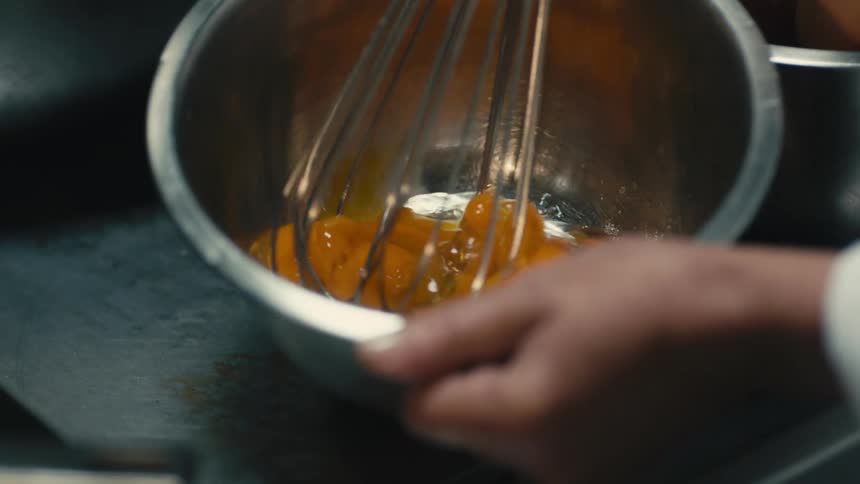 A hand is holding a silver bowl with eggs inside and a wire whisk is stirring them