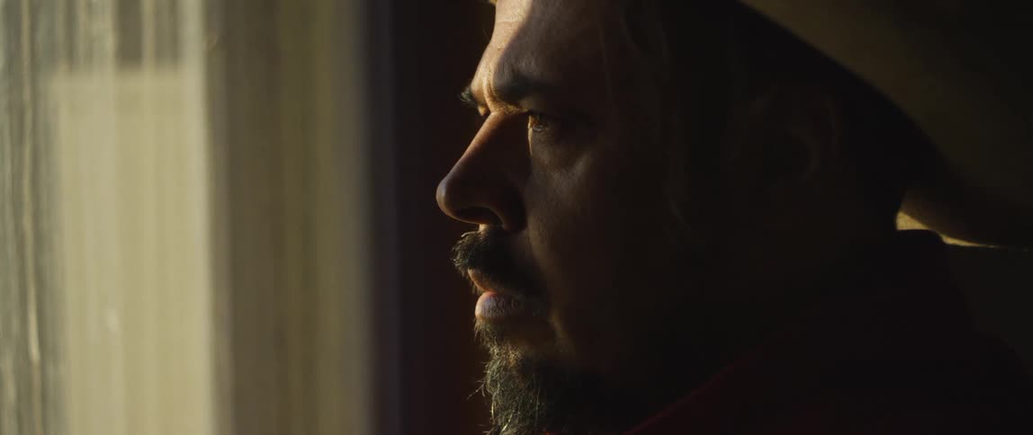 Man stands staring out window through thin curtains as sunlight and shadow cover his face