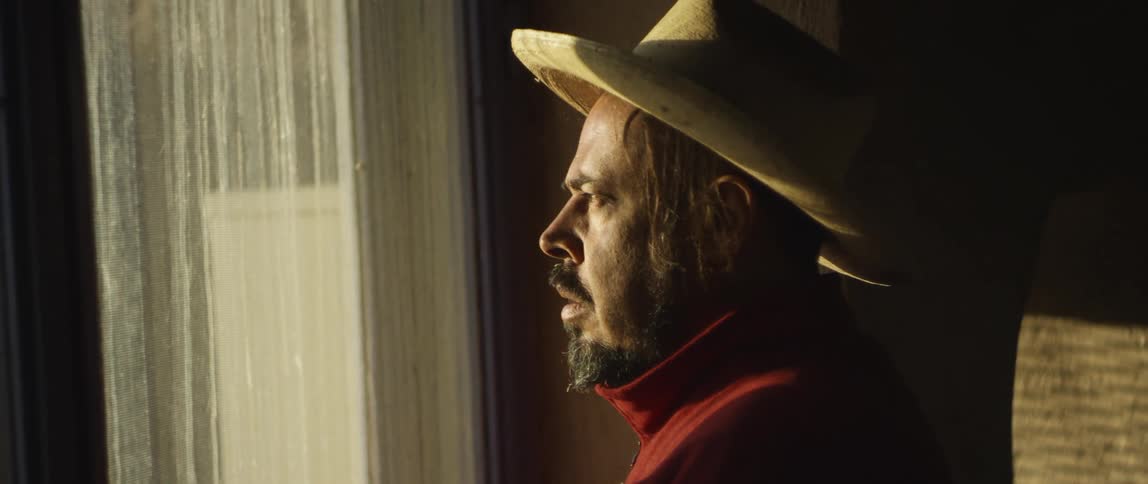 Anxious man with cowboy hat beard and red shirt looks out the window