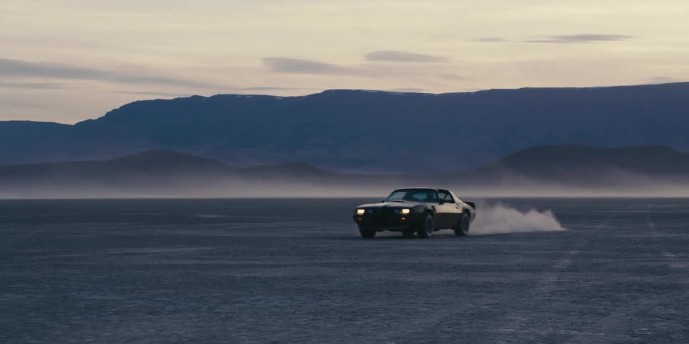 A lone car on a desert plain takes off leaving a trail of dust in the early morning with mountains in the background