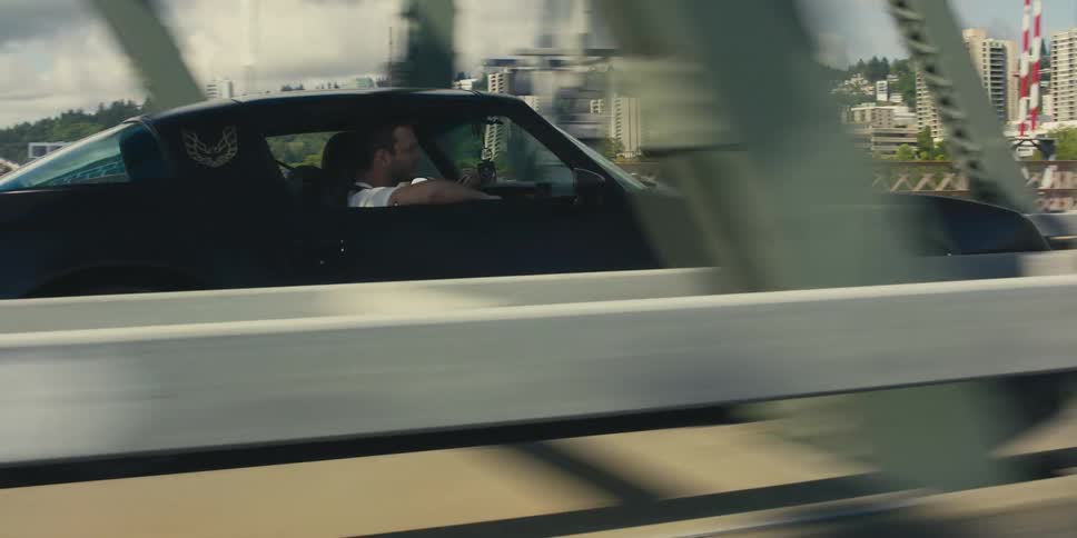 A man rides in the passenger seat of a sports car with his arm leaning on the window as the cross a bridge