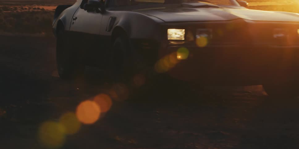 A black car with headlights on is traveling on a road as the sun sets