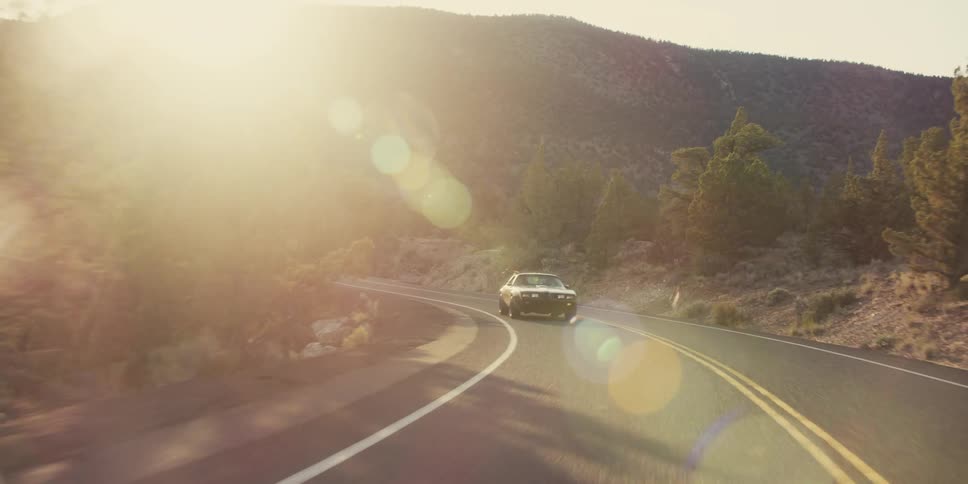 A sports car is traveling along a winding mountain road with trees in the evening