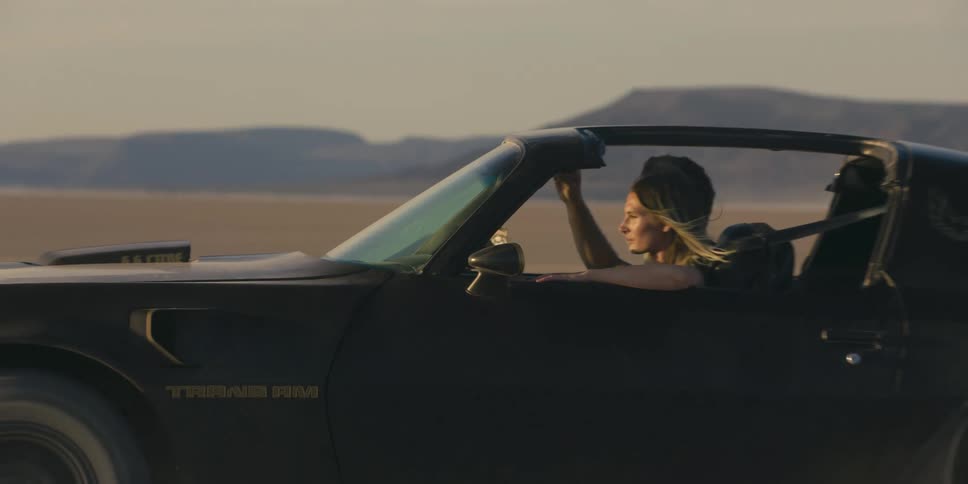 Man and woman speed across desert with top down kicking up sand with mountains on horizon