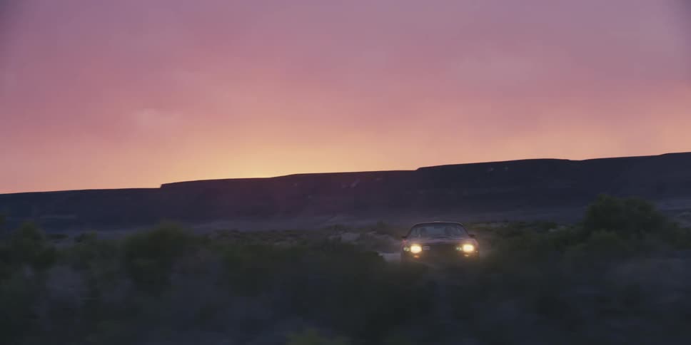A car driving on a desert with a mountain range the sky and clouds in the background