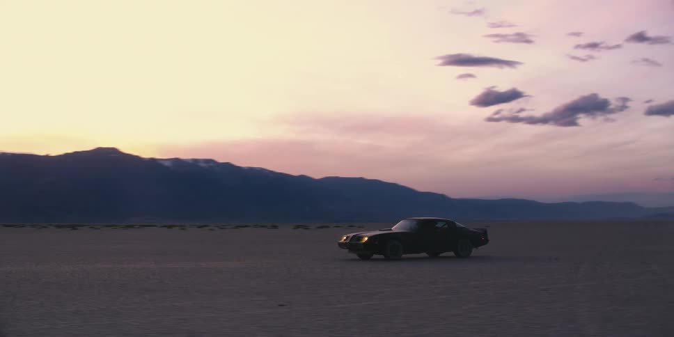 Man gets into sports car parked in desert surrounded by mountains under a red sky and drives off