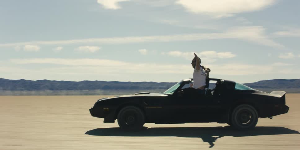 Young couple stand out of a moving black muscle car in the desert
