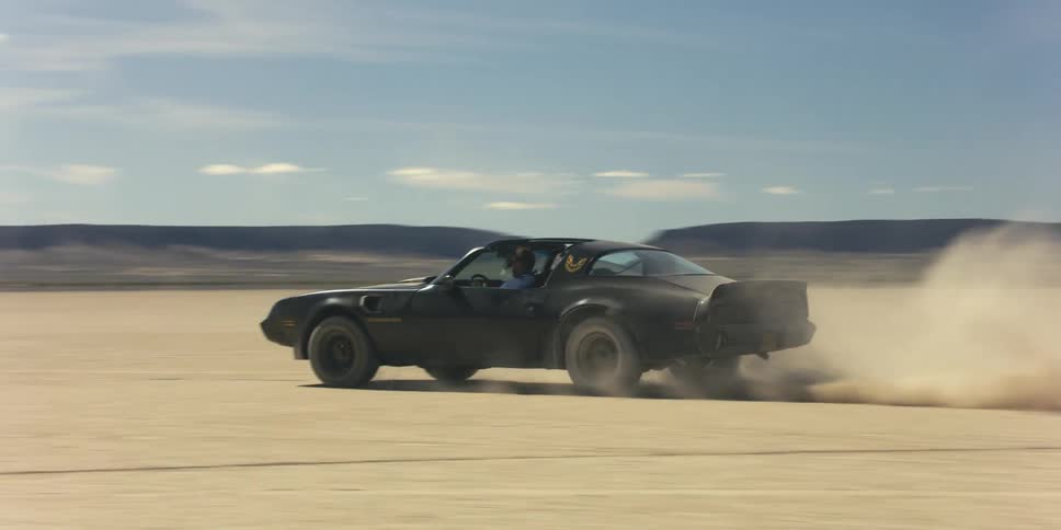 A car driving on a desert with a mountain range the sky and clouds in the background