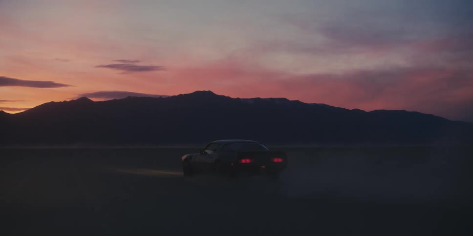 A car driving on a desert with a mountain range the sky and clouds in the background