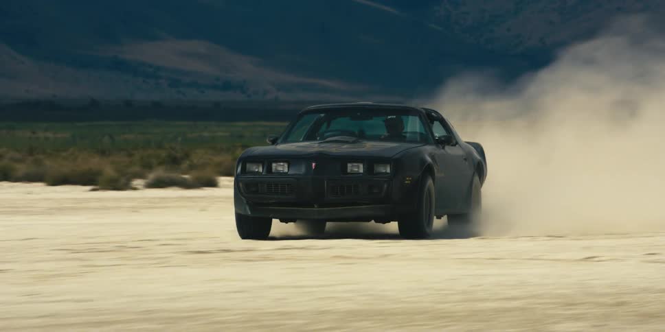 Man drives car along sand past green field with mountains in background