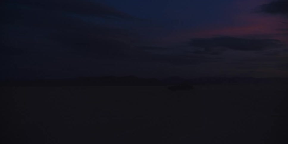 Car drives across desert under red sky with mountain in background and into darkness of storm