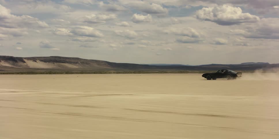 Sports car speeds across desert kicking up dust with mountains on two sides in background under cloudy sky