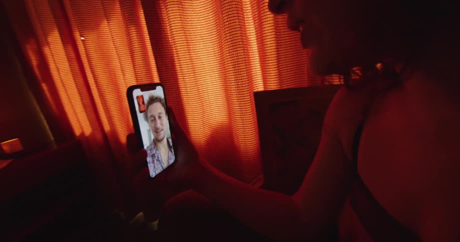 Woman with wavy hair and black undershirt makes a video call on smartphone in dim lit room