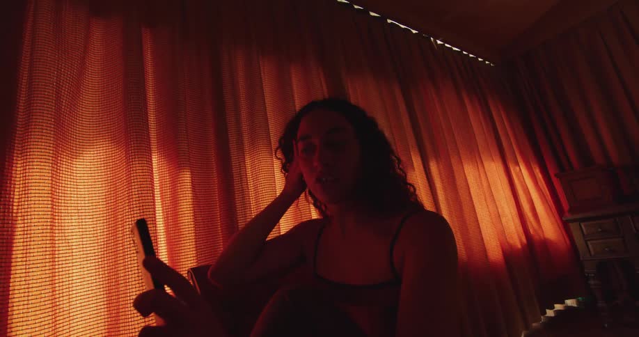 A woman is standing inside a home near orange curtains and plays with her hair as she has a video chat on her cell phone