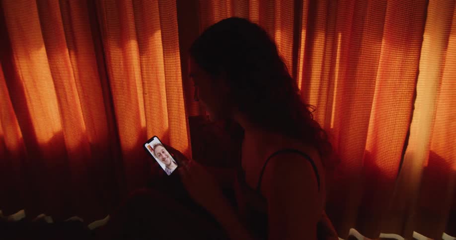 A woman is standing inside a home near orange curtains and having a video chat on her cell phone