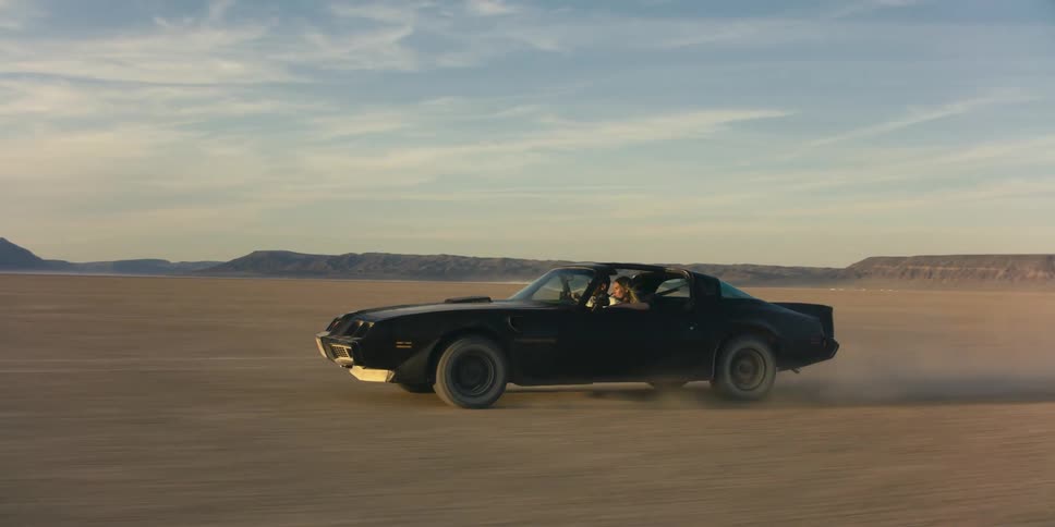 Couple driving a black muscle car on the desert near the mountains at sunset