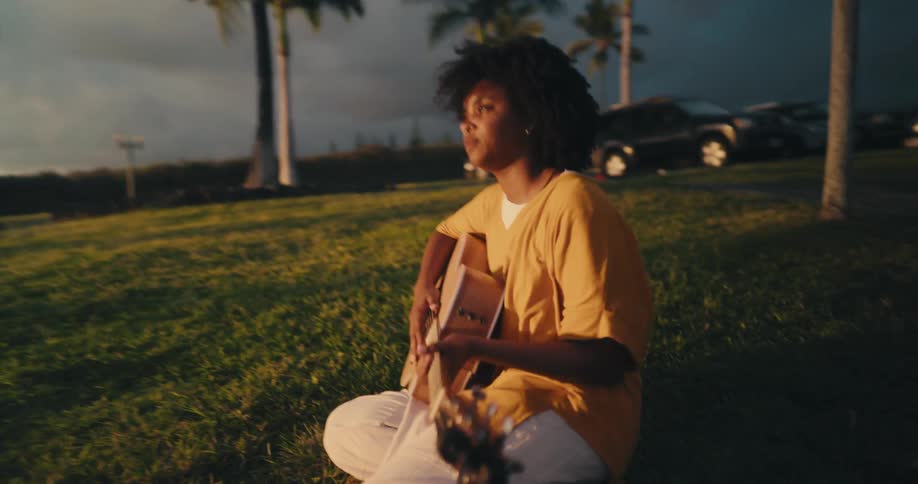 A woman sitting on grass playing a guitar with the sky and clouds in the background