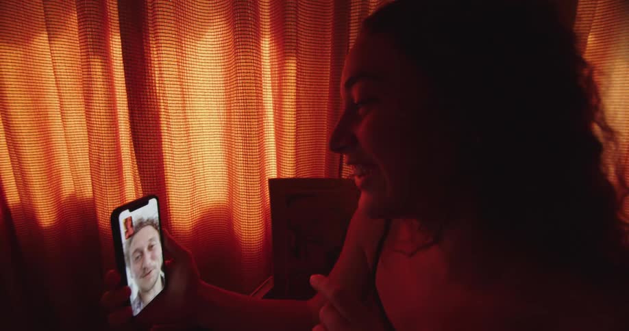 A woman in a room video chatting on a smart phone with a window and a wall in the background