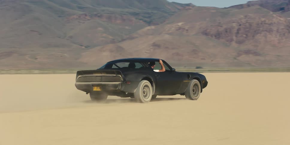A black trans am with ttops is traveling across a desert with mountains in the distance