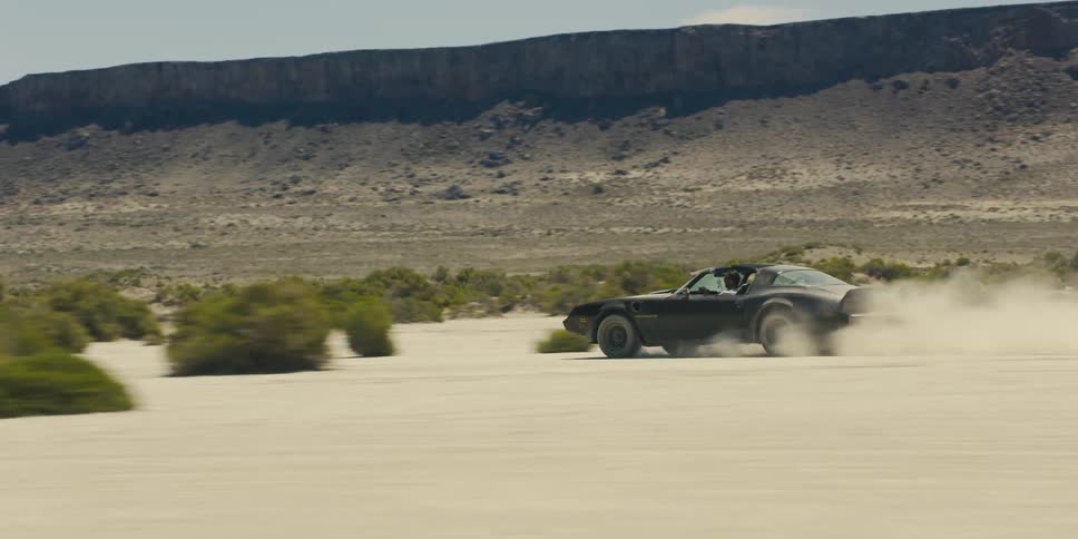 People speed along desert scrub in sports car with top down kicking up sand with mountains in background