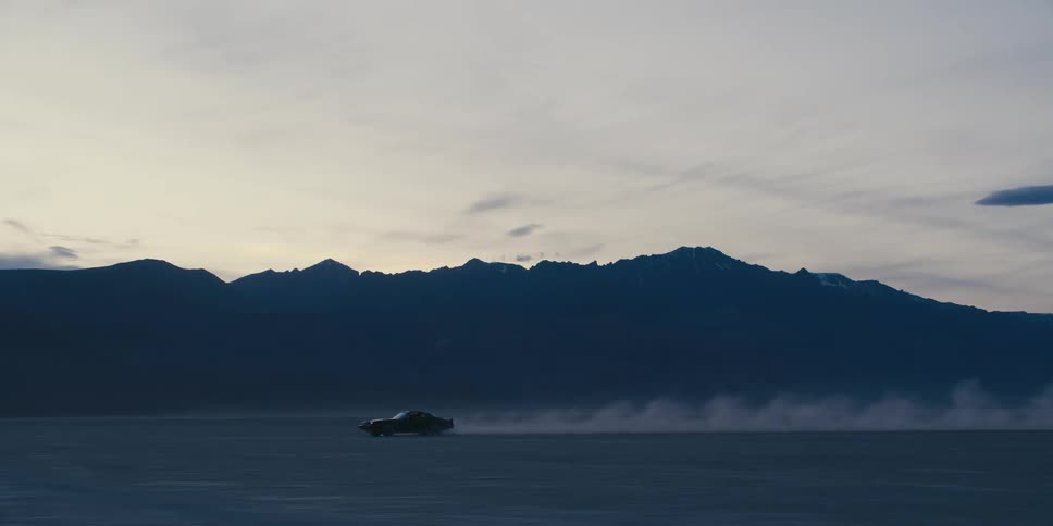 A black trans am with ttops is traveling across a desert with mountains in the distance