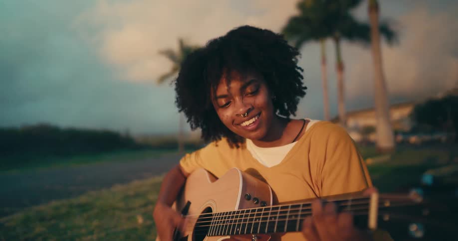Young woman palying acoustic guitar while sitting in the grass near palm trees at sunset