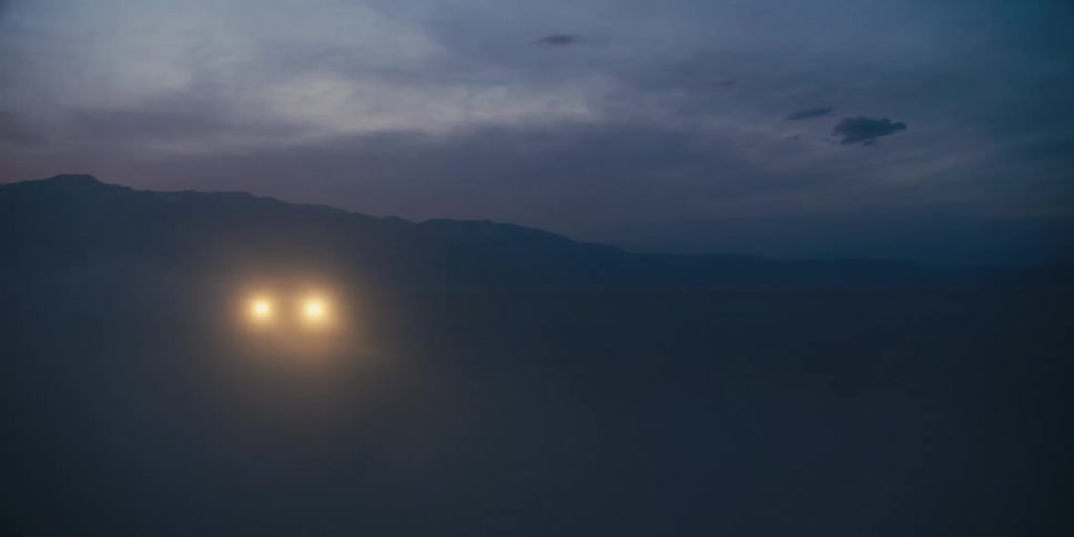 A car driving on a desert with a mountain range the sky and clouds in the background