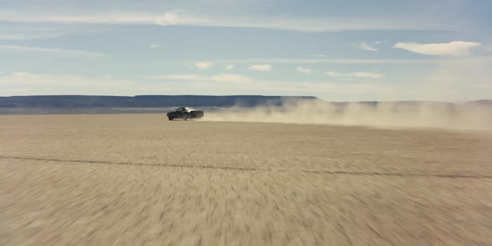 Sports car races across desert plain kicking up dust with mountains on horizon