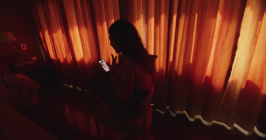 A woman is standing inside a home near orange curtains and having a video chat on her cell phone