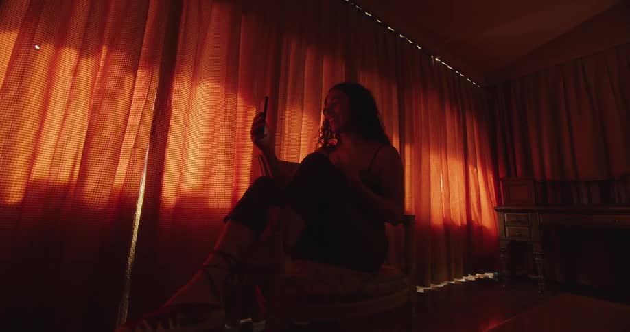 A woman in a room sitting on a chair looking at a smart phone with a wall and window in the background