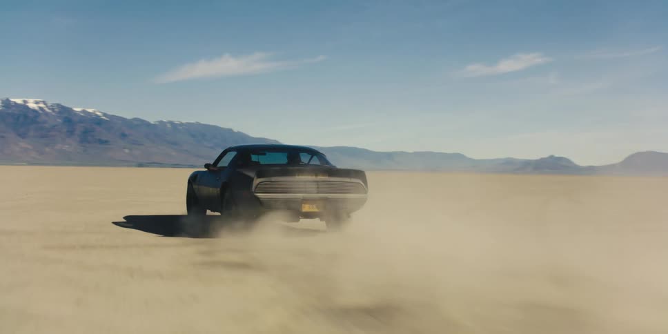 A car driving on a desert with a mountain range the sky and clouds in the background