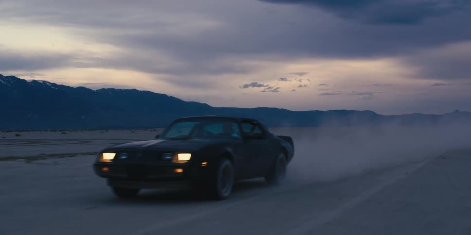 Car drives across desert sands creating dust cloud with mountain in background under cloudy sky
