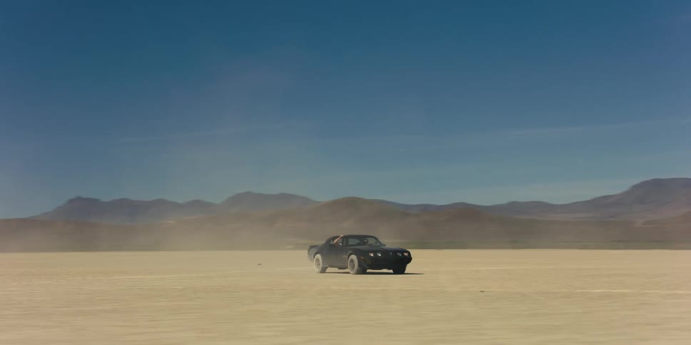A man driving a sports car offroad through the desert stops the vehicle to exit the vehicle and switches seats with a woman who promptly drives off