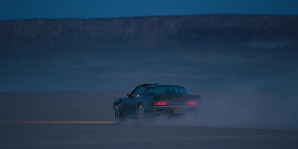 A car driving on a desert with a mountain range the sky and clouds in the background