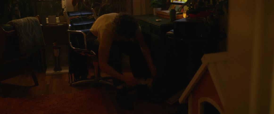 A teenage boy is sitting in a chair in front of a computer and he bends down to take off his shoes